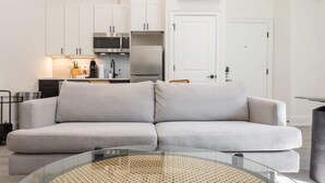 Ein modernes Wohnzimmer mit einem grauen Ecksofa, einem Glastisch und einer Küche im Hintergrund.