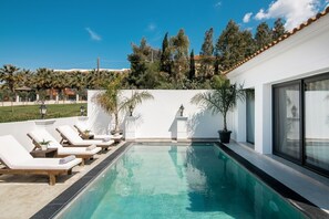 Una piscina moderna al aire libre con sillones de descanso, una pared blanca y vistas a árboles y edificios al fondo.