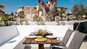 Un espace de restauration en plein air avec une table en bois dressée pour deux personnes, entourée de chaises, et offrant une vue sur des bâtiments et des palmiers en arrière-plan.