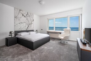 Une chambre à coucher moderne, dotée d’un grand lit, d’un bureau et d’une chaise, et offrant une vue sur la mer grâce à une grande fenêtre.