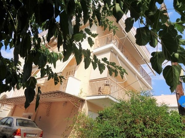 A multi-story building with balconies, a car parked in front, and surrounding greenery.