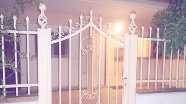 Een wit metalen hekwerk met decoratieve details, dat leidt naar een gebouw met een beige muur en een raam.