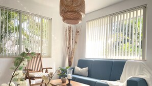 Una sala de estar con un sofá azul, una silla de madera y una mesa de centro. Hay dos ventanas con persianas verticales y una lámpara de techo tejida sobre la mesa.