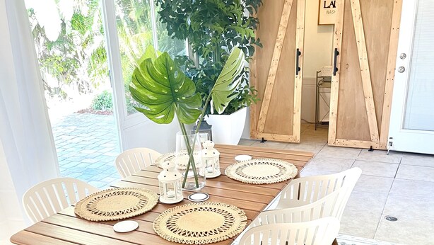 Una mesa de comedor de madera con individuales tejidos, sillas de plástico blanco y una gran planta como centro de mesa.
