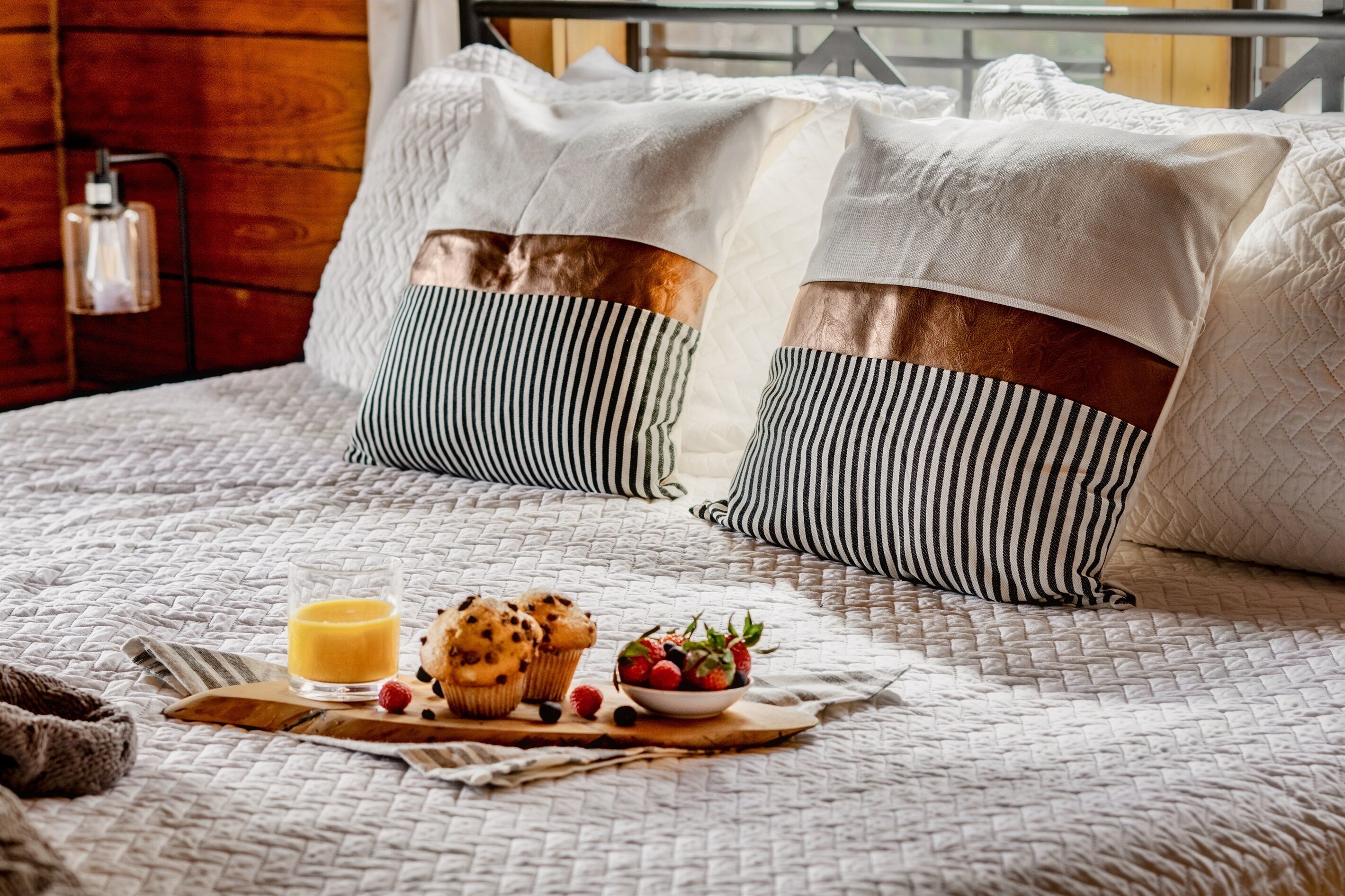 A neatly made bed with striped pillows, a glass of orange juice, muffins, and a bowl of fruit on a wooden tray.