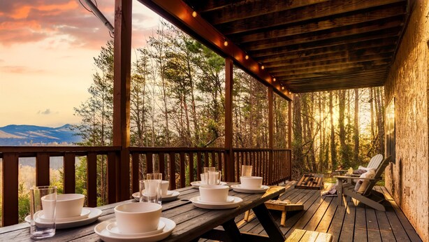A wooden deck with a table set for dining, overlooking a forested area with mountains in the distance. - Stunning VIEWS, Luxury, Golf, Hot Tub, Arcade, Pet Friendly & Nearby Attractions (Sevierville)