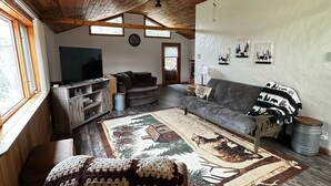 Ett mysigt vardagsrum med ett högt träpanelat tak, en stor platt-TV, en grå soffa, en mönstrad matta och ett fönster med utsikt över träd.