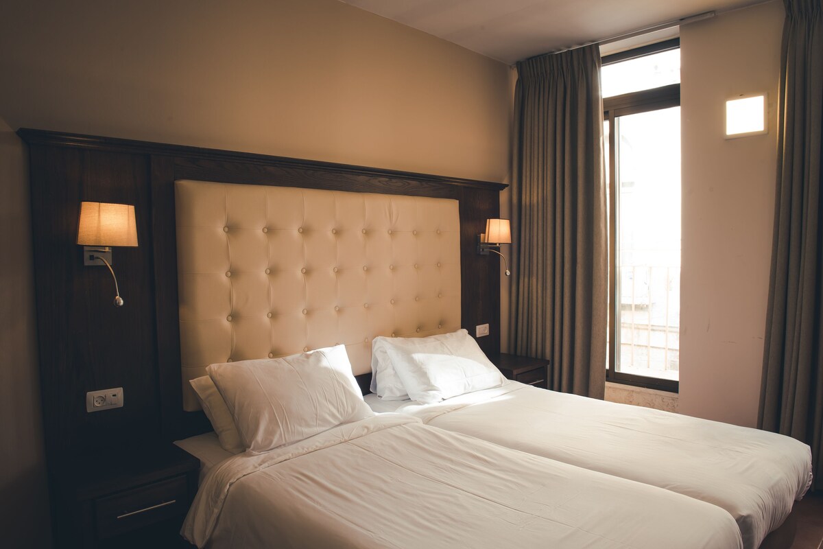 A hotel room with a large bed, two bedside lamps, a window with curtains, and a view of the outside.