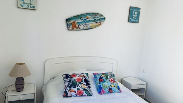 Een slaapkamer met een wit bed, twee bloemenkussens, een nachtkastje met een lamp en twee wanddecoraties.