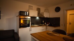 A compact kitchen with a microwave, oven, and sink. There are cabinets, a clock, and a dining area with a table and chairs.