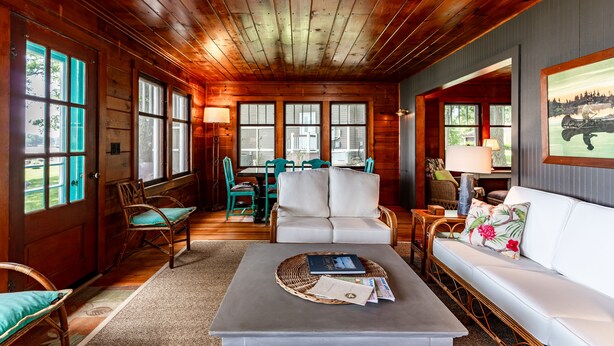 L’intérieur chaleureux d’un chalet avec des poutres apparentes, un canapé blanc, des fauteuils en osier et un coin repas comprenant une table et des chaises.