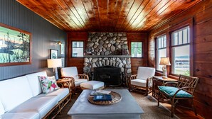 Un intérieur de chalet chaleureux avec une cheminée en pierre, un plafond en bois, des meubles blancs et de grandes fenêtres.