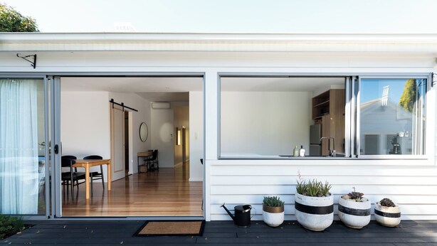 A modern house with a sliding glass door leading to a wooden interior, a balcony with potted plants, and a clear view of the sky.