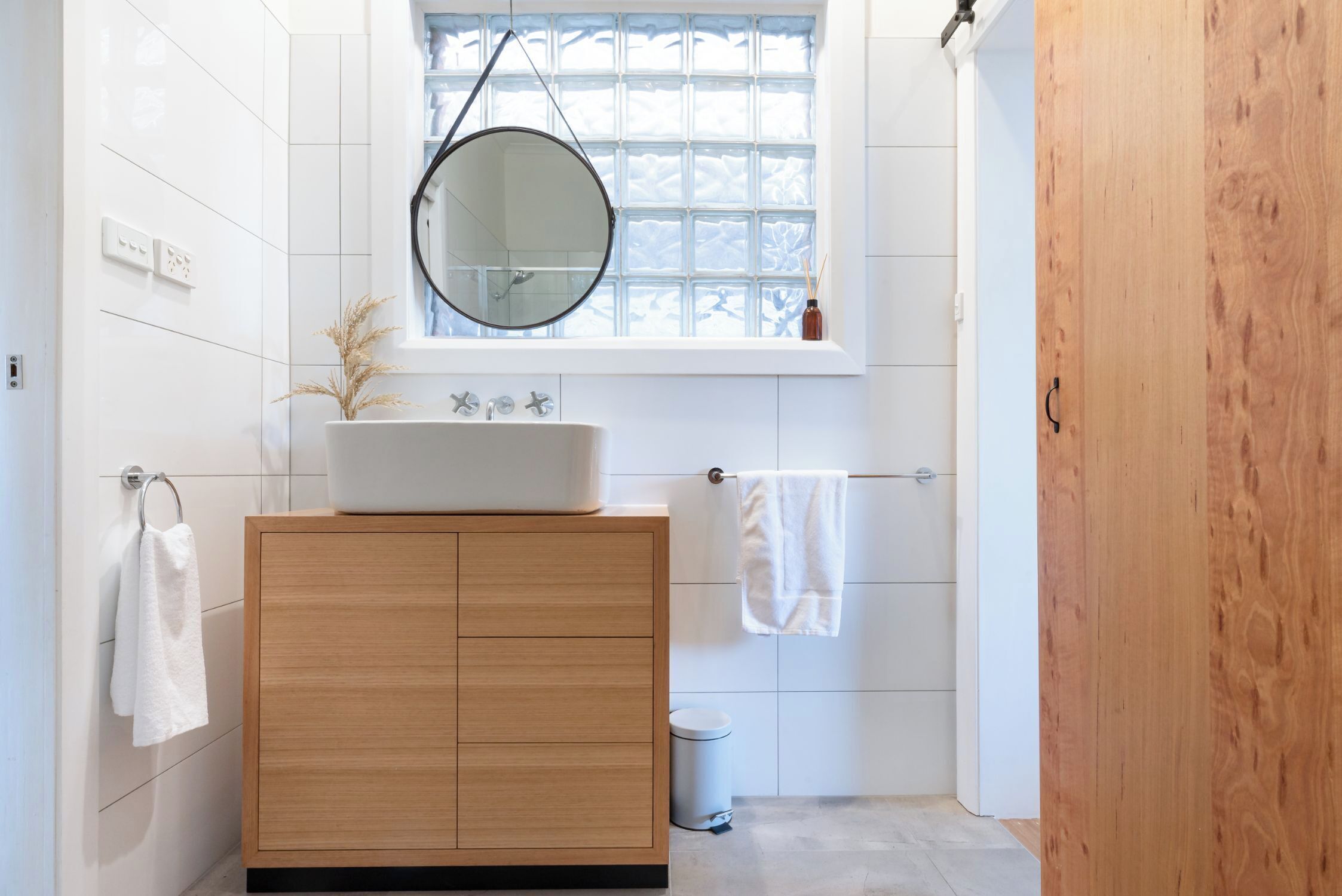 Une salle de bain moderne avec un meuble-lavabo en bois, un miroir rond, une fenêtre et une petite poubelle.