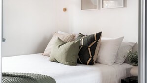 A neatly made bed with a white comforter and several pillows, a small bedside table with a plant, and a wall-mounted picture frame. - Walter Cottage: As Cosy as it is Convenient (South Launceston)