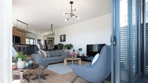 A modern living room with a grey sofa, a coffee table, and a TV. There is a dining area with a wooden table and chairs, and a kitchen in the background. - Impressively Spacious, Modern & Convenient (Newstead)