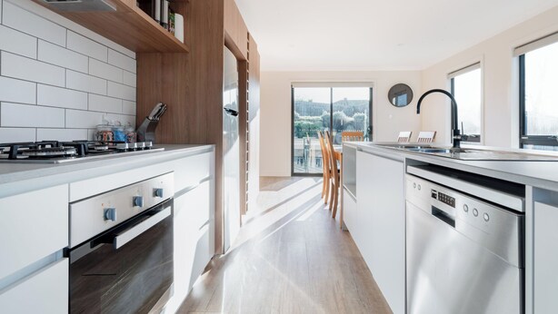 A modern kitchen with white cabinetry, a built-in oven, and a dishwasher. There is a wooden shelf above the stove and a large window providing a view of the outdoors. - Modern & Convenient in Newstead (Newstead)