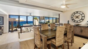 A spacious dining area with a large wooden table and wicker chairs, a large wall clock, and a view of the ocean through large windows. - Hawaii Life Rentals Presents Puu Poa 302 (Princeville)