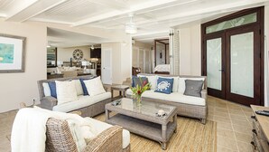 A spacious living room with wicker furniture, a coffee table with decorative items, and a view of the kitchen and dining area. - Hawaii Life Rentals Presents Puu Poa 302 (Princeville)