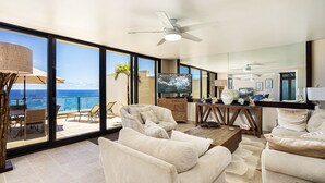 A spacious living room with a large TV, comfortable seating, and a balcony with a view of the ocean. - Hawaii Life Rentals Presents Puu Poa 302 (Princeville)