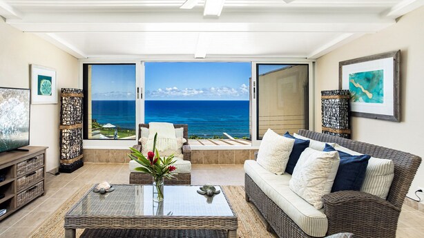 A living room with a view of the ocean, wicker furniture, and a television. - Hawaii Life Rentals Presents Puu Poa 302 (Princeville)