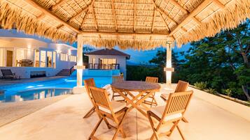 A poolside area with a thatched roof, wooden chairs, and a table.