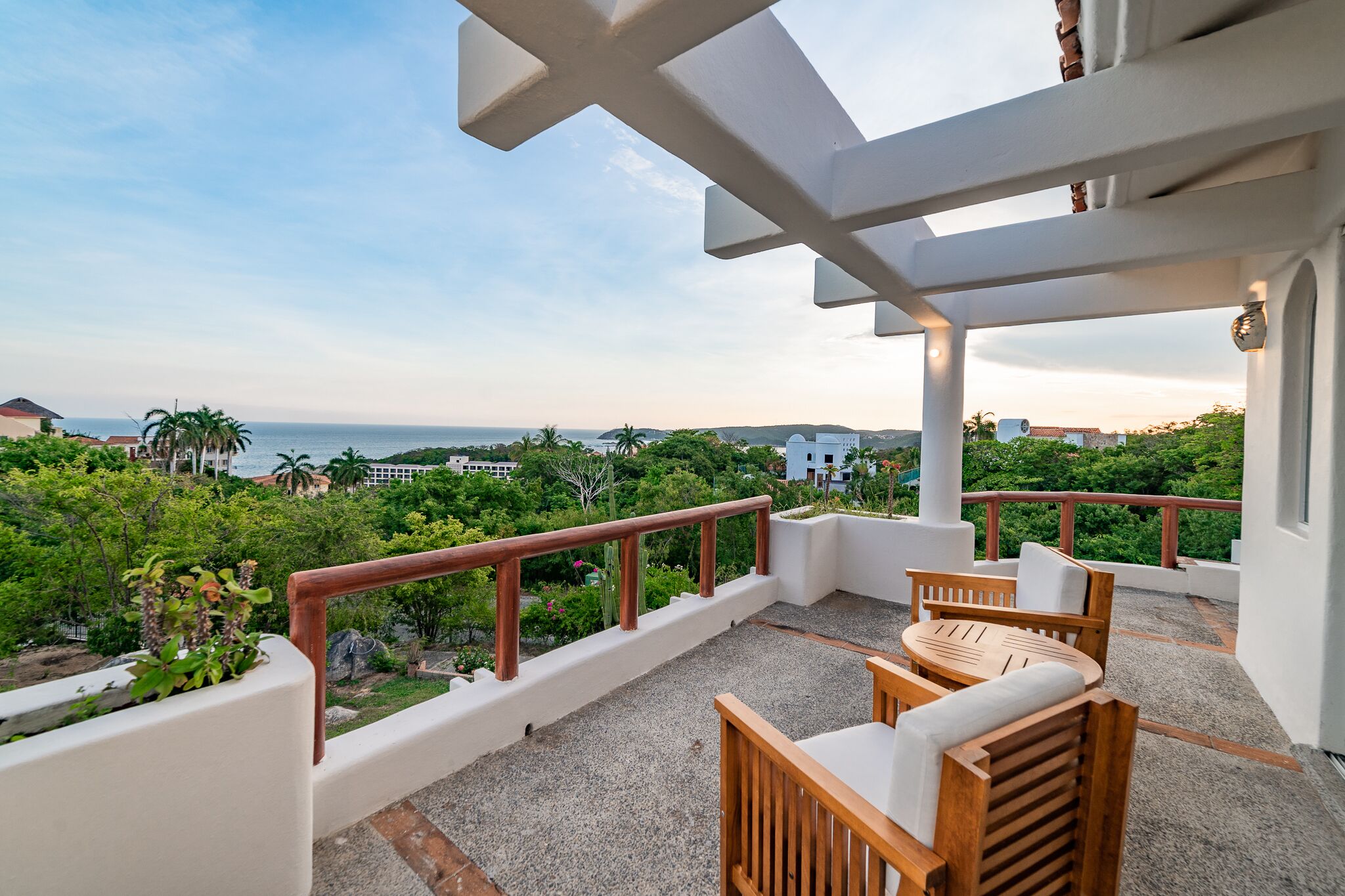 A balcony with a view of the sea, surrounded by greenery and white walls.