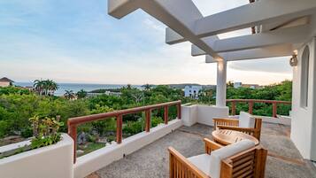 A balcony with a view of the sea, surrounded by greenery and white walls.