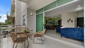 Un apartamento moderno con balcón, sillas de mimbre, un sofá azul y una cocina visible a través de un vidrio.