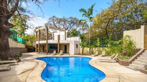 Una piscina con borde curvo, rodeada por una solana de hormigón. Hay sillones de descanso y una escalera que lleva a un nivel superior.