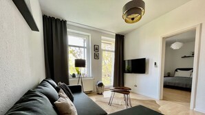 Un salon moderne avec un canapé vert foncé, une petite table d’appoint en bois, un lampadaire et un téléviseur à écran plat fixé au mur.