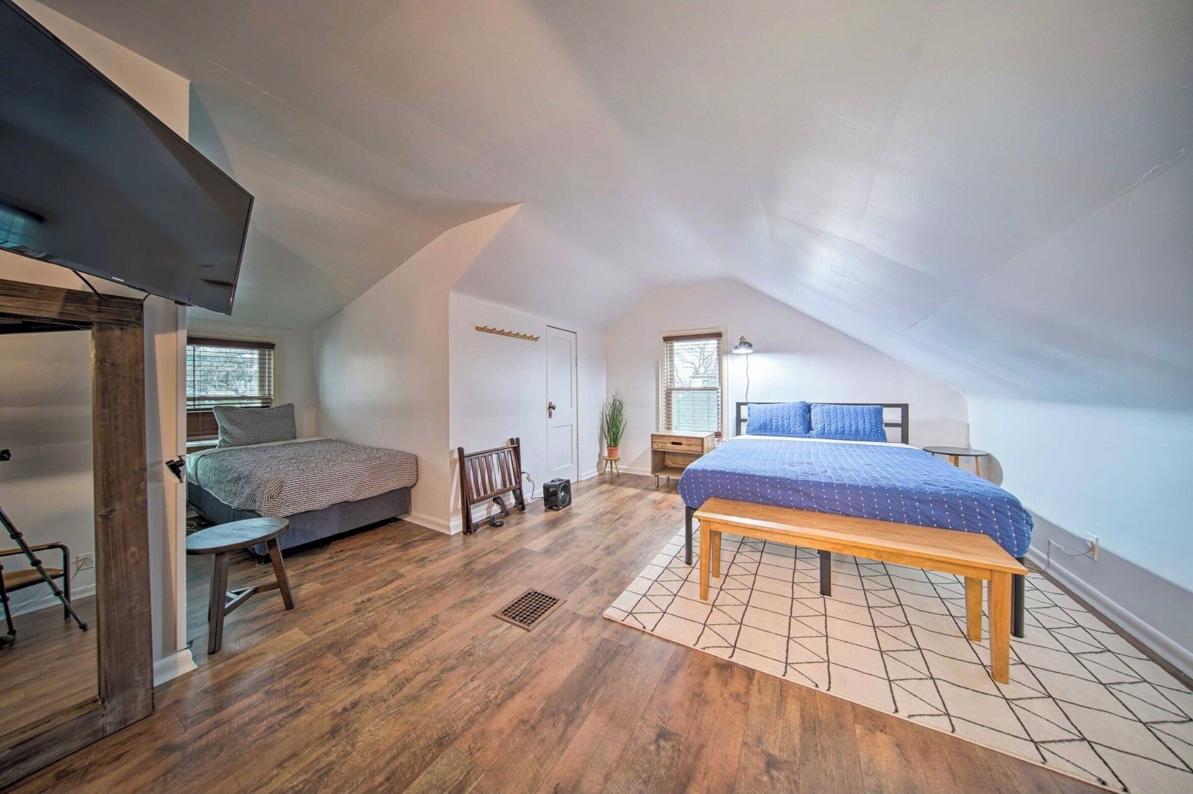 A bedroom with a sloped ceiling, a bed with a blue bedspread, a wooden bench, a small table, and a radiator.