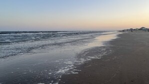 A beach with a calm sea, a sandy shore, and a few buildings in the distance. - 3min walk to beach,hot tub, b-ball, pet friendly, parking for RV’s (Bolivar Peninsula)