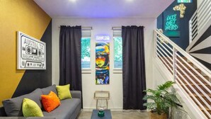 Ein modernes Wohnzimmer mit einem grauen Sofa, farbenfrohen Kissen, einem schwarzen Beistelltisch, einem großen abstrakten Gemälde und einer Neonleuchtreklame an der Wand.
