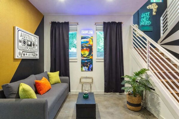 A modern living room with a grey sofa, colorful cushions, a black side table, a large abstract painting, and a neon sign on the wall.