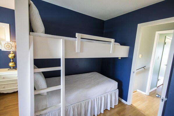 Uma cama beliche num quarto com paredes azul-escuras, uma cômoda de madeira e uma porta que dá para outro espaço.