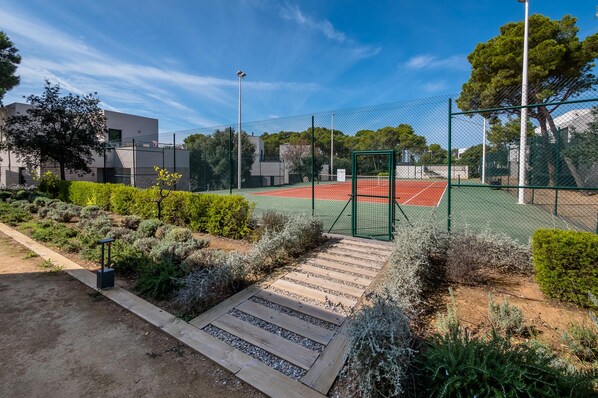Un court de tennis entouré d’une clôture verte, bordé d’arbustes et d’arbres aménagés.