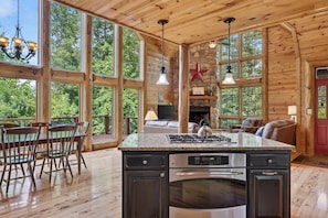A spacious kitchen with a large island, wooden walls, and large windows offering a view of the outdoors. - River Woods Retreat: Hot Tub, Hikes, 2 Fireplaces, EV (Bentonville)