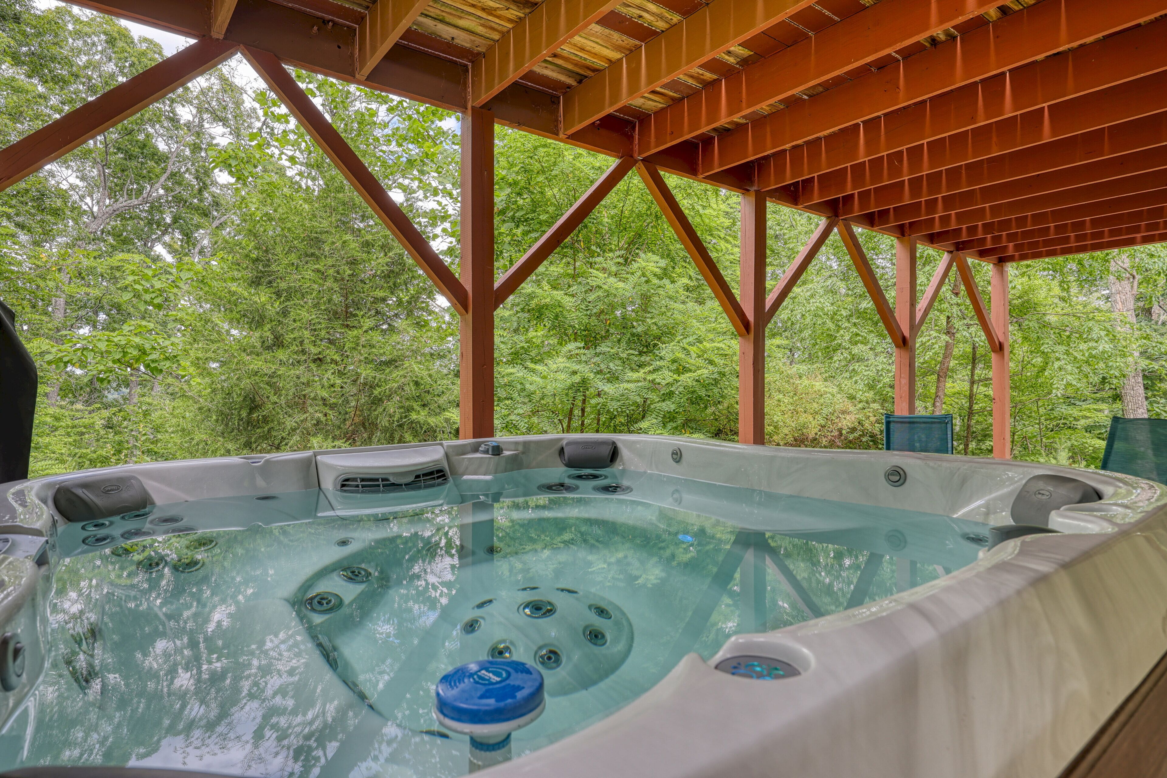 A hot tub under a wooden structure with a forest backdrop.