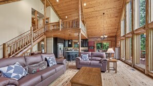 Interior espacioso de una cabaña con escalera de madera, sala de estar con sofás de cuero, una mesa de centro de madera y ventanas grandes.