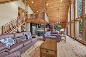 A spacious cabin interior with a wooden staircase, a living area with leather sofas, a wooden coffee table, and large windows. - River Woods Retreat: Hot Tub, Hikes, 2 Fireplaces, EV (Bentonville)