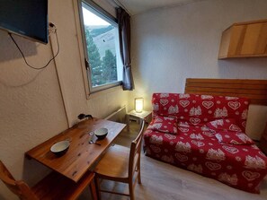 Ein kleiner Raum mit einem Holztisch, einem roten, gemusterten Stuhl, einer einzelnen Nachttischlampe und einem Fenster mit Blick auf eine gebirgige Landschaft.