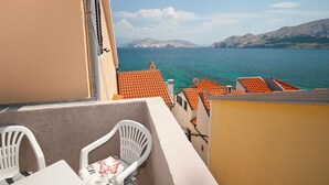Een balkon met uitzicht op zee en bergen, witte plastic stoelen en terracotta dakpannen.