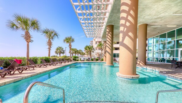 A swimming pool with clear blue water, surrounded by lounge chairs and palm trees, under a shaded pergola. - Crescent Keyes PH18-2BR/2BA Oceanfront-Pools (North Myrtle Beach)