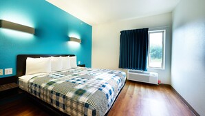 A hotel room with a bed, a window with curtains, a wall-mounted air conditioner, and a blue accent wall.