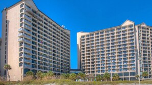 Two high-rise hotels with multiple floors and balconies, located near a beach with sparse vegetation.