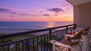 A balcony with a view of the ocean at sunset, featuring wooden chairs and two glasses of red wine.