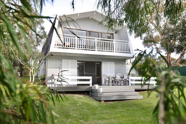 A two-story house with a balcony, a patio with outdoor furniture, and a well-maintained lawn. - Sabi@Robe - Garden Retreat - 300m from Long Beach (Robe)