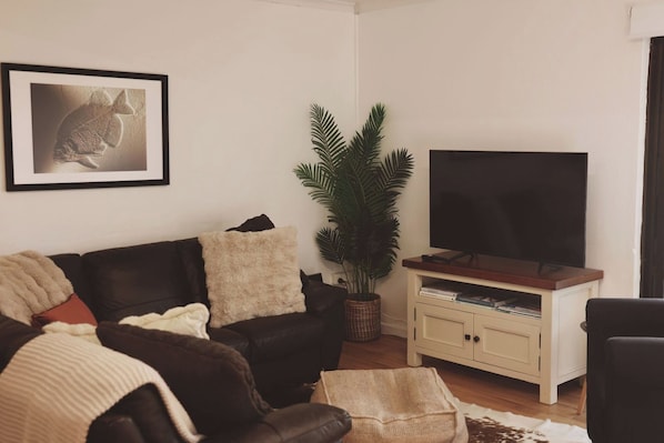 A living room with a black leather sofa, a television on a wooden stand, a potted plant, and a framed picture on the wall. - Sabi@Robe - Garden Retreat - 300m from Long Beach (Robe)
