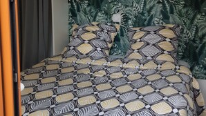 A bed with a patterned bedspread, a headboard with a leaf pattern, and a wooden shelf above the bed. - Bungalow aux Villages Sous les Pins, à 10 Minutes des Plages (LEON)